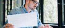 Serious pensive thoughtful focused young casual business accountant bookkeeper in office looking at and working with laptop and income tax return papers and documents