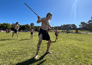 A group of Indigenous performers showcasing a traditional dance with spears at an outdoor event on a sunny day.